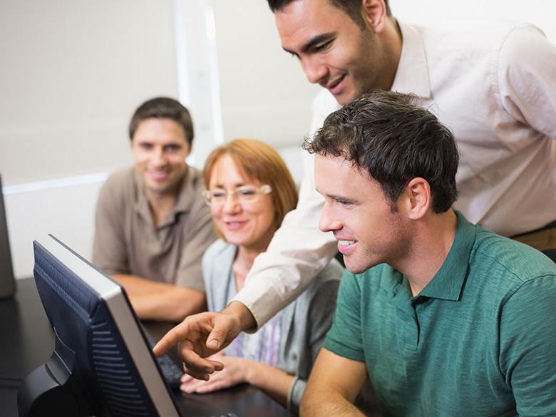 Adult students sit at computers and are assisted by a teacher